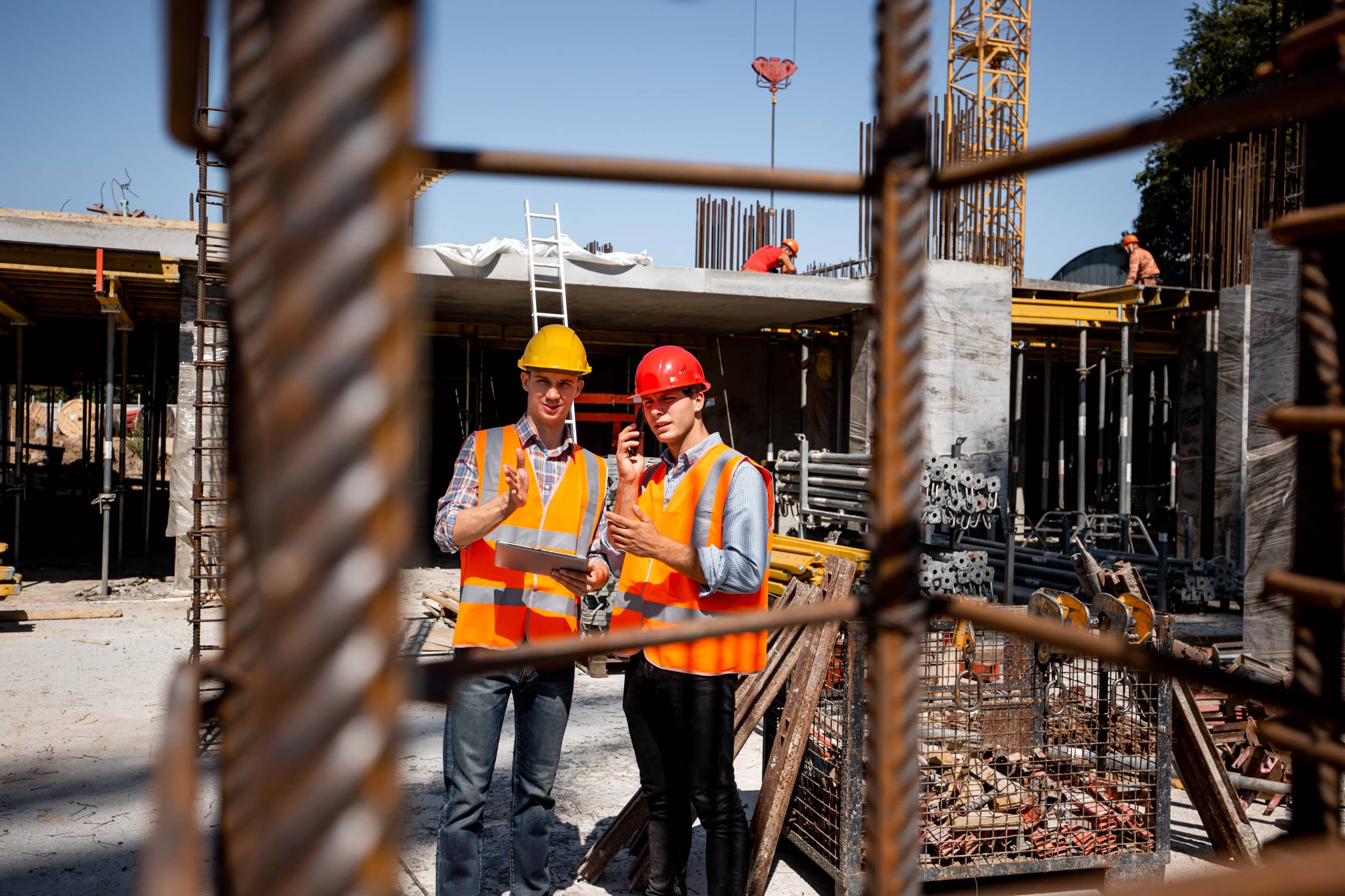 Architect and structural engineer in orange work vests and helmets discuss a building project on the open air building site with a lot of steel frames