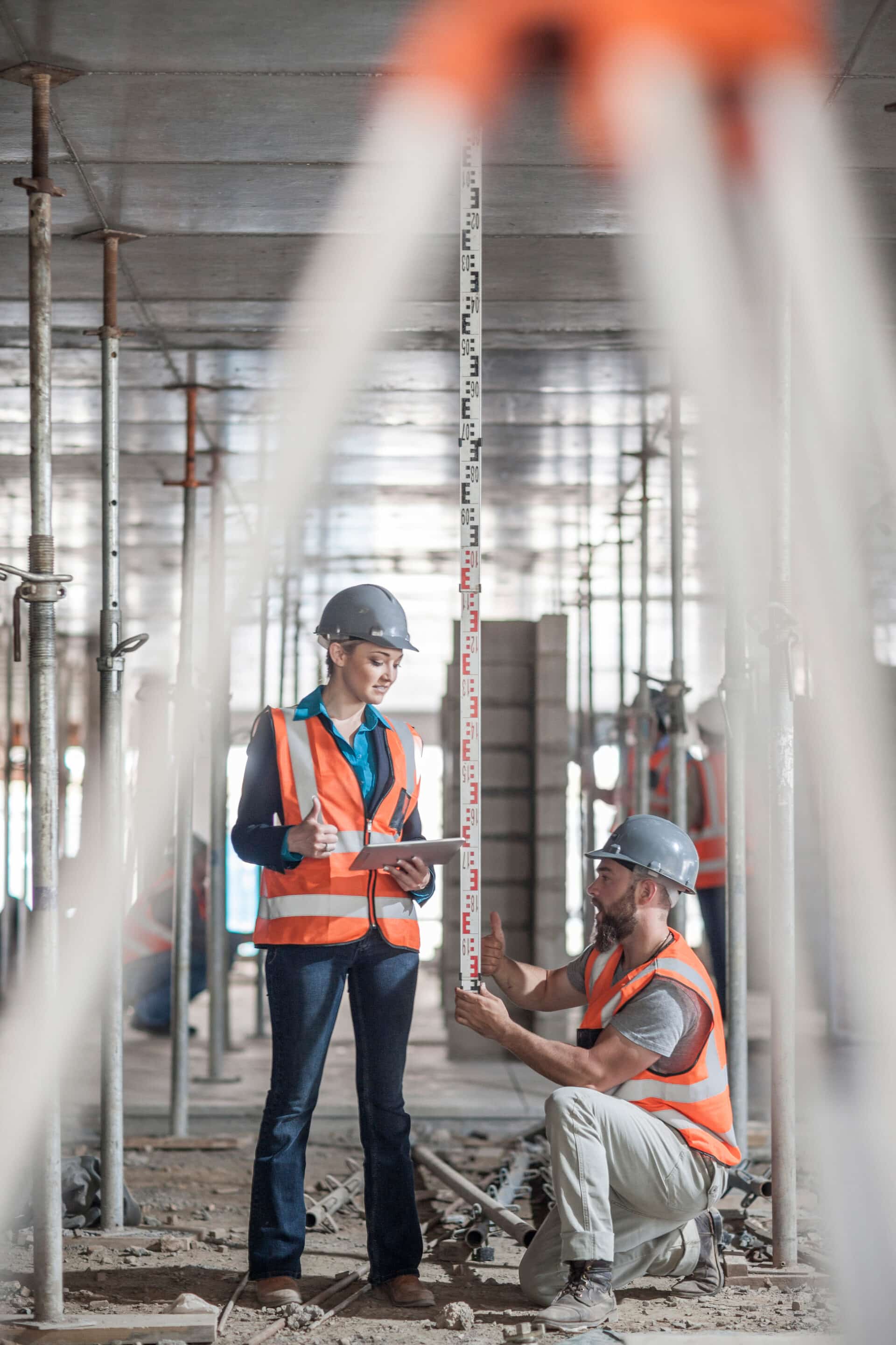 Male and female builders measuring construction with ruler Deux professionnels du bâtiment en gilets et casques de sécurité effectuent une mesure de hauteur avec une mire à l’intérieur d’un bâtiment en construction.